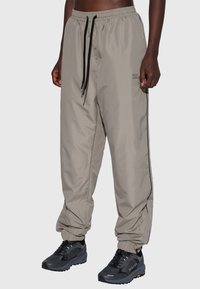 Ljusbeige, löst sittande byxor med elastisk midjeband, svart dragsko och sidodetaljer, med en logotyp tryckt på låret.