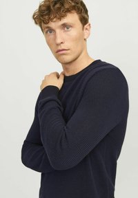 Navy blue knitted sweater featuring a textured pattern, crew neckline, and ribbed cuffs, worn by a model with curly hair.