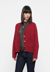 Rød kabelstrikket cardigan med V-hals, gullknapper og en frontlomme, båret over en svart bluse med prikker og mørke jeans.
