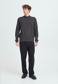 Grijze gebreide trui met een hoge hals, gecombineerd met zwarte broek en zwarte schoenen. De textuur lijkt glad en de pasvorm is ontspannen.