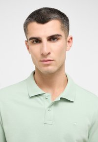Šviesiai žalia polo marškinėliai iš tekstūruoto audinio, su dviejų mygtukų apykakle ir mažuoju logotipu kairėje krūtinės pusėje.