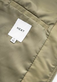 Étiquette de vêtement blanche sur tissu beige avec le nom de la marque NEXT et la taille S fabriqué en Chine, visible sur l'étiquette.