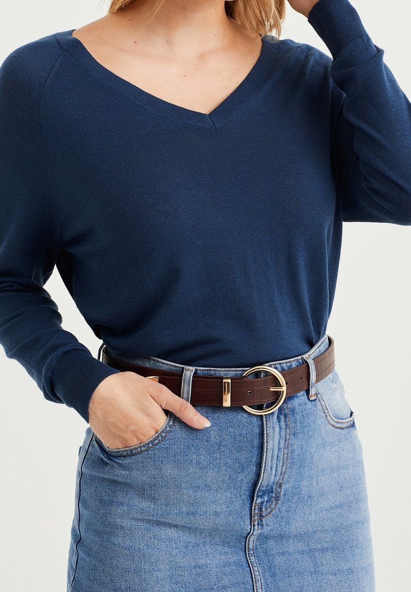 Femme portant un pull bleu marine à col en V rentré dans une jupe en denim bleu clair taille haute avec une ceinture marron et une boucle ronde dorée.