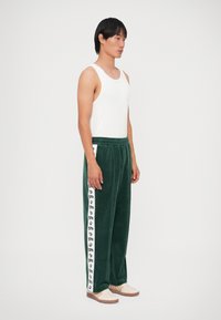 Pantalons de survêtement en velours vert avec des bandes latérales blanches arborant des logos noirs, associés à un débardeur blanc et des chaussures beiges. Coupe droite.
