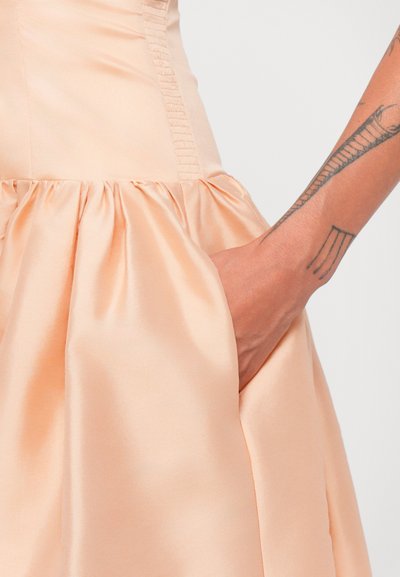 Persikkavärinen satiinik dress, jossa on tiukka yläosa, rypytetty hame ja sivutasku. Dressissä on hieno kiilto ja teksturoitu sivusaumaviimeistely.
