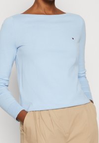 Maglione azzurro chiaro con scollo a barca, maniche lunghe, polsini a costine e un piccolo logo sul petto. Realizzato in un materiale morbido e testurizzato.
