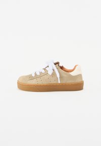 Beige mockasinsneakers med vita skosnören, perforerade detaljer på sidan och en brun gummisula. Klacken har en kontrasterande krämfärgad accent.