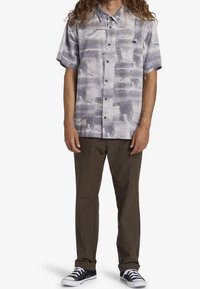 Camicia a maniche corte con bottoni di colore grigio chiaro con motivi astratti, abbinata a pantaloni verde oliva e sneakers nere.