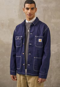 Mörkblå denimjacka med vit sömnad, med fyra framfickor och guldfärgad hårdvara. Bärs över en grå tröja.