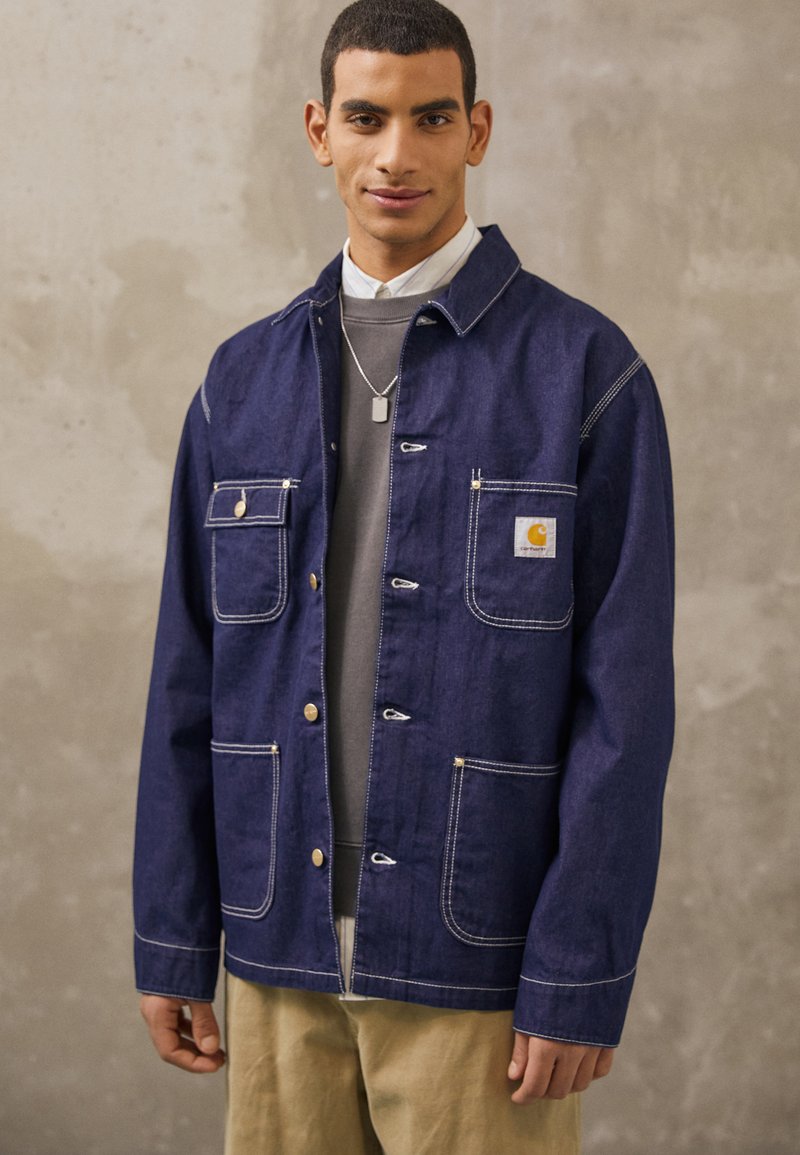 Mörkblå denimjacka med vit sömnad, med fyra framfickor och guldfärgad hårdvara. Bärs över en grå tröja.