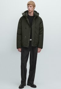 Giacca imbottita verde scuro con cappuccio, dotata di chiusura con zip, due tasche frontali e una vestibilità rilassata, indossata sopra un maglione nero e pantaloni grigi.