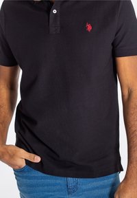 Svart pikétröja i bomull, med en röd logotyp på bröstet, korta ärmar och krage med två knappar. Bärs tillsammans med blå jeans.