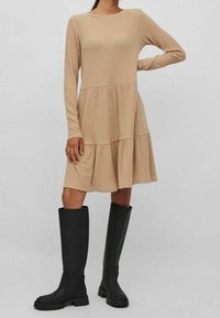 Robe beige à manches longues en maille côtelée avec un ourlet à volants, associée à des bottes en caoutchouc noires montantes avec semelles épaisses et texture lisse.