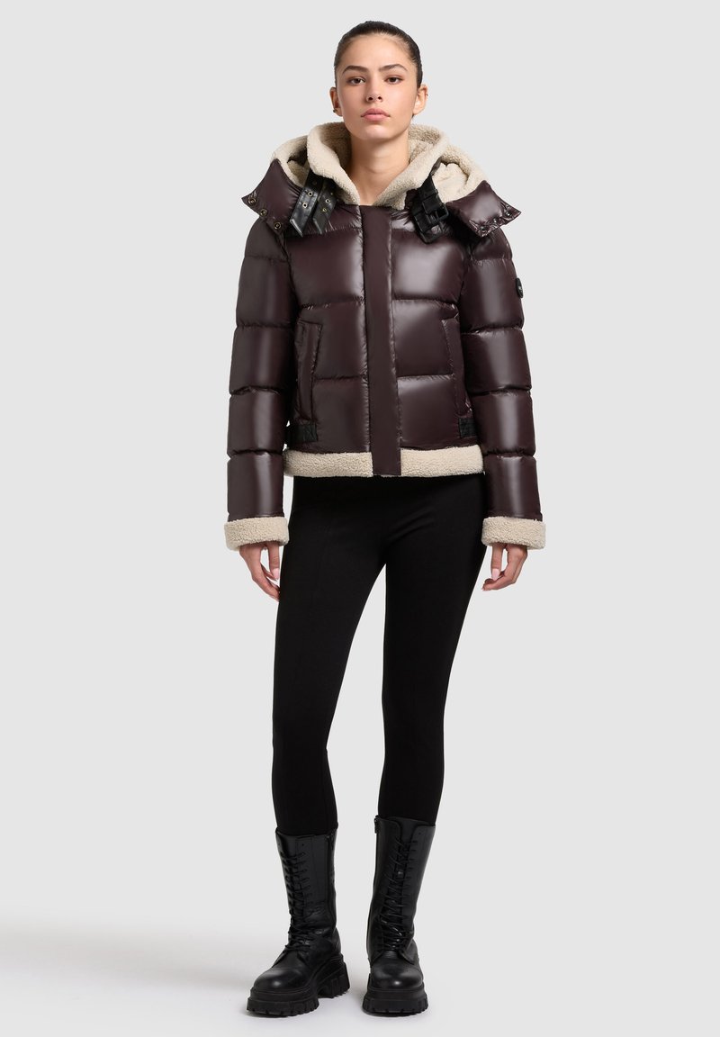 Giacca puffer marrone scuro con cappuccio foderato in pile, dettagli neri e polsini in shearling. Il design presenta una vestibilità cropped e una forma squadrata.