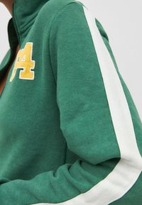 Giacca verde con zip, strisce bianche sulle spalle e sulle maniche e toppa gialla "24" sul petto, indossata da una persona.