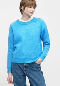 Jaunas žmogus su trumpais rudiems plaukais, vilkintis ryškiai mėlynu megztiniu ir mėlynomis džinsomis, su viena ranka kišenėje, žiūrintis į šoną.