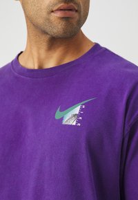 Uomo che indossa una maglietta Nike viola con il logo swoosh turchese e una grafica di un pallone da basket sul petto, mostrato dalla nuca fino a metà torso.