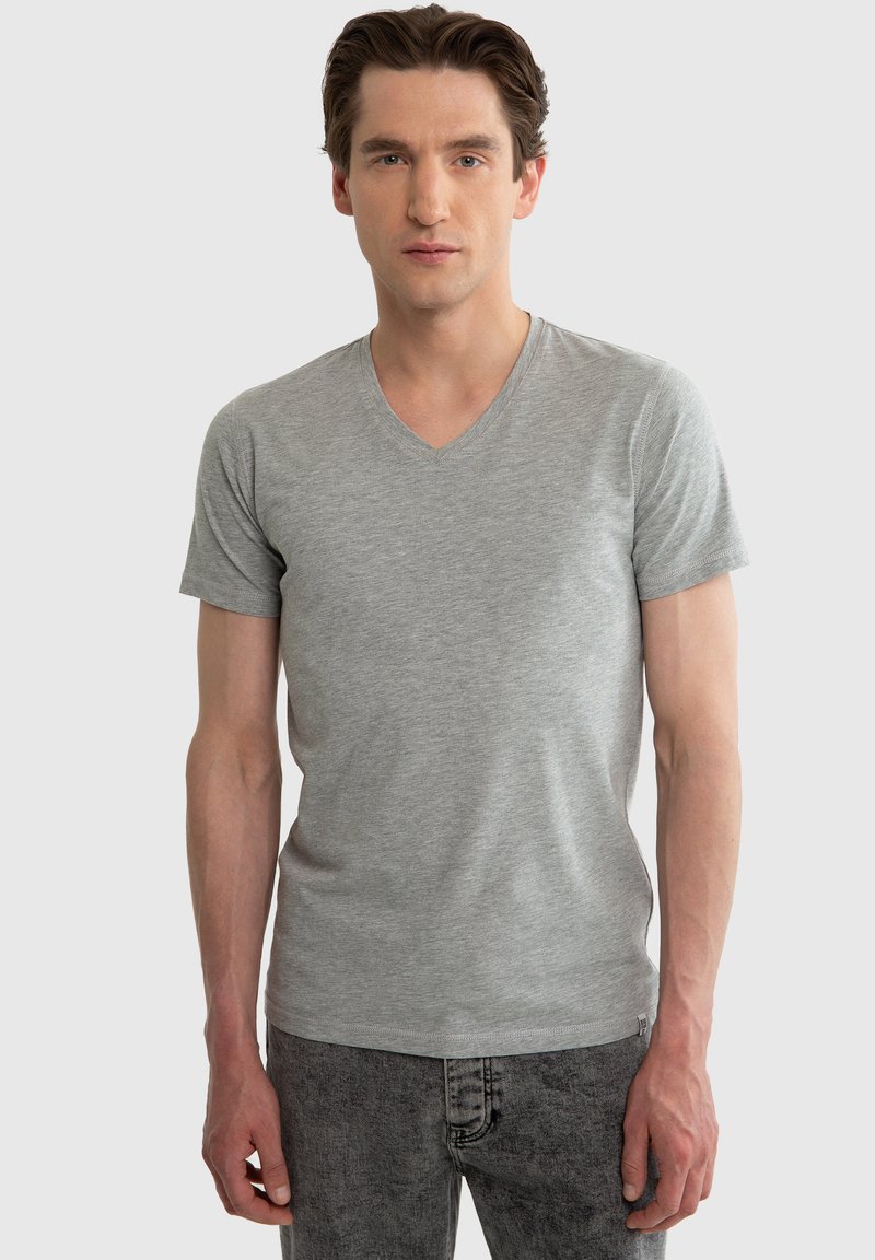 Jonge man met kort bruin haar, gekleed in een lichtgrijs T-shirt met V-hals en donkergrijze spijkerbroek, staand tegen een effen achtergrond.