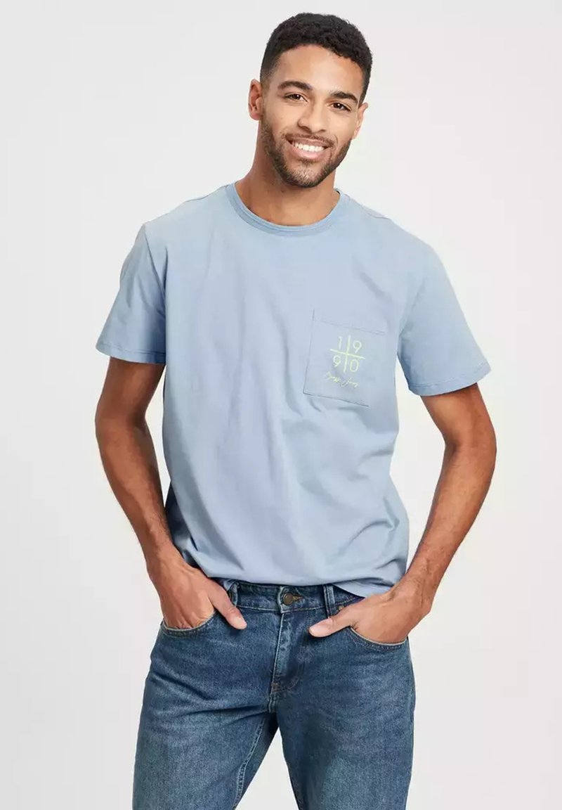 Jasnoniebieska koszulka z krótkim rękawem z kieszonką na piersi, z zielonymi graficznymi napisami. Noszona z niebieskimi dżinsami, ma luźny krój i miękką teksturę.