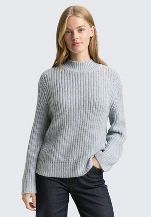 Junge Frau mit langen blonden Haaren, die einen hellgrauen gerippten Strickpullover und dunkle Jeans trägt, steht mit einer Hand in der Tasche.