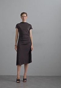 Brown, mid-length dress with short sleeves, featuring ruched side detailing. Smooth fabric with a fitted silhouette, paired with open-toe black shoes.
