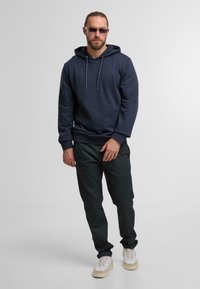 Donker marineblauwe hoodie met gestreepte trekkoorden, gecombineerd met zwarte textuurbroek en lichte sneakers, staand tegen een effen achtergrond.