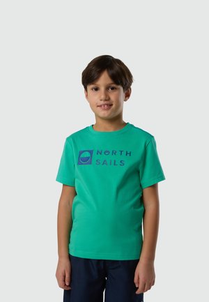 Ragazzo con capelli corti e scuri che indossa una maglietta verde "North Sails" e pantaloncini blu scuro, in piedi contro uno sfondo chiaro e uniforme.
