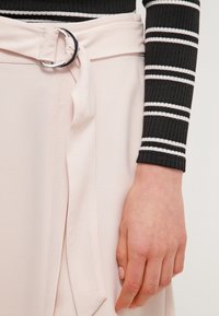 Jupe enveloppante rose pâle avec une boucle circulaire argentée et un détail de lien, associée à un haut à manches longues rayé noir. Tissu texturé.