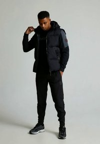 Svart puffervest med hette, grå detaljer og glidelåslukking, kombinert med svarte joggebukser. Modellen har på svarte joggesko og en mørk skjorte.