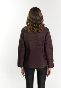 Un giubbotto imbottito di un viola scuro uniforme con un motivo trapuntato e una vestibilità rilassata, indossato su pantaloni neri. Cuciture in evidenza sugli maniche.
