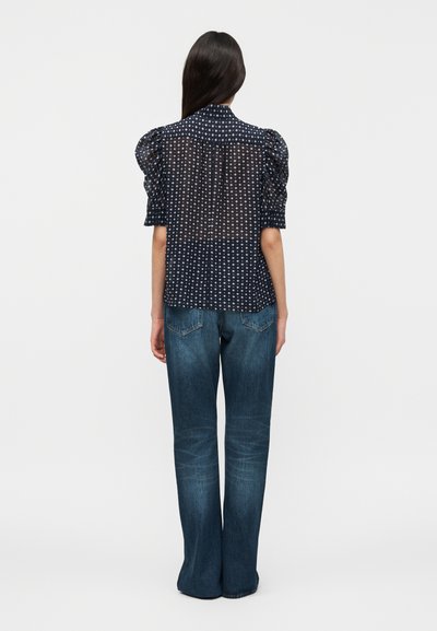 Γυναίκα που στέκεται με την πλάτη, φορώντας μπλε σκούρα διάφανη μπλούζα με λευκά πουά και φουσκωτά μανίκια, συνδυασμένη με μπλε τζιν, σε μονόχρωμο φόντο.