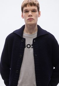 Tengerészkék kardigán texturált kötött mintával, sálgallérral és fekete gombokkal, világosszürke pulóver fölött, melyen élénk logó található.