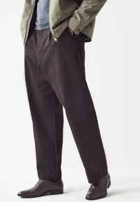 Donkerbruine taps toelopende broek van zacht materiaal, met zijzakken en een gladde textuur, gecombineerd met strakke bruine nette schoenen.