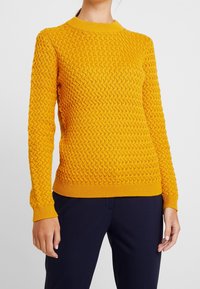 Tröja i stickat i klar gult, med ett texturerat mönster och rundad hals. Ribbad ärm- och fållkant, med en figurnära silhuett för en åtsittande passform.