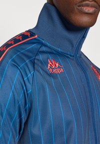 Μπλε ζακέτα με φερμουάρ, με κάθετες ρίγες και κόκκινες λεπτομέρειες. Διαθέτει λογότυπο Kappa στο στήθ�ος και στον ώμο, κατασκευασμένη από λείο, ελαφρύ υλικό.