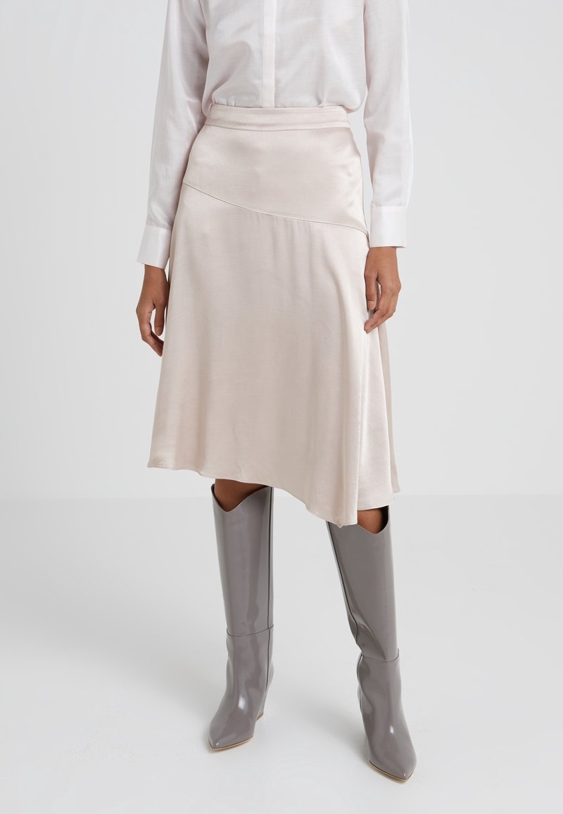 Femme portant une jupe midi en satin rose pâle avec un ourlet asymétrique et des bottes grises pointues montant jusqu'au genou, associées à une chemise blanche à manches longues.