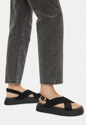 Sandalias de ante negro con tiras entrecruzadas, un acento de hebilla y una suela negra gruesa y texturizada. Combinadas con pantalones de mezclilla cortos gris.