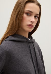 Felpa grigia con cappuccio in tessuto morbido e finitura testurizzata. Presenta lacci al cappuccio e una vestibilità rilassata, vista accorciata.