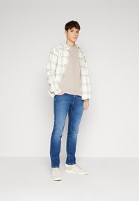 Chaqueta de cuadros blanca sobre una camiseta beige, jeans azules y zapatillas beige con rayas blancas. Pose de pie sobre un fondo neutro.