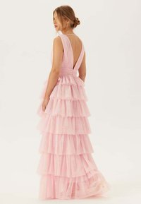 Puchata różowa sukienka z warstwowymi, falbaniastymi segmentami. Bez rękawów, z głębokim dekoltem na plecach w kształcie litery V. Wykonana z lekkiego, przezroczystego materiału o płynącej sylwetce.