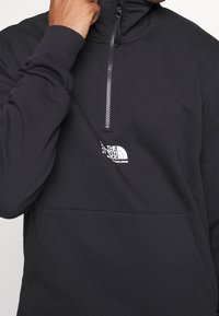 Personne portant un pull noir à demi-fermeture éclair avec poche kangourou et logo blanc The North Face sur la poitrine, main près du col.