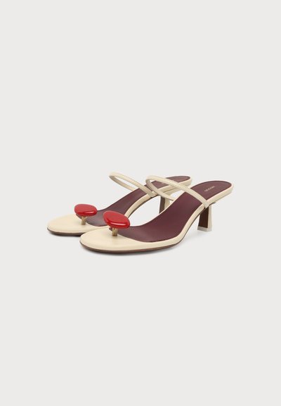 Paire de sandales à talons crème avec fines brides, semelles bordeaux et accents rouges circulaires audacieux à l'avant, sur fond blanc uni.