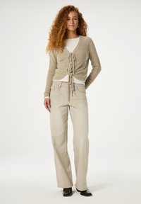 Beige gebreide cardigan met een strikdetail aan de voorkant, gedragen over een aansluitend wit topje, gecombineerd met lichtbeige wijde broek en zwarte schoenen.