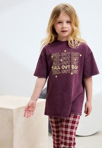 T-shirt violet en coton avec inscription "Fall Out Boy" en jaune, coupe ample, manches courtes, assorti à un pantalon à carreaux bordeaux et beige.