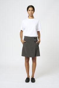 Grå omslagskjol med två svarta knappar och sidofickor, i kombination med en enkel vit t-shirt och svarta båtbygda skor, stående mot en neutral bakgrund.