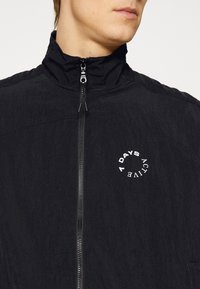 Chaqueta negra con cremallera y cuello alto. Presenta una tela texturizada y un logo circular blanco que dice "7 DAYS ACTIVE" en el lado izquierdo.