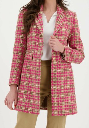 Femme portant un long manteau rose et beige à motif pied-de-poule par-dessus un chemisier blanc et un pantalon beige, visible du cou jusqu'à la mi-cuisse.