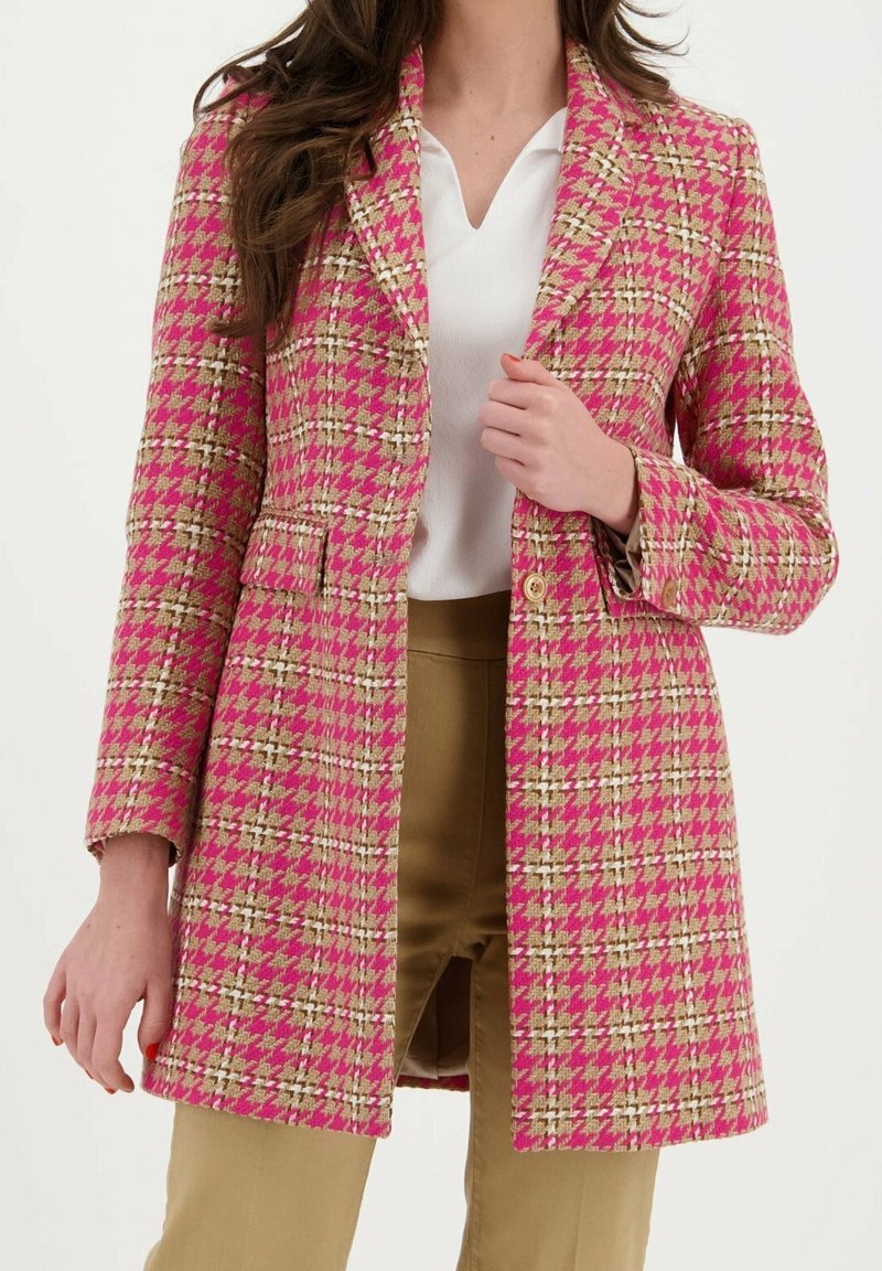 Femme portant un long manteau rose et beige à motif pied-de-poule par-dessus un chemisier blanc et un pantalon beige, visible du cou jusqu'à la mi-cuisse.