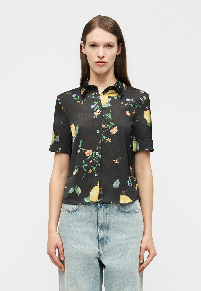 Γυναίκα που φοράει μαύρο πουκάμισο με κοντά μανίκια και κίτρινα και πορτοκαλί λουλουδάτα σχέδια, συνδυασμένο με ανοιχτόχρωμα τζιν ψηλόμεσα παντελόνια.