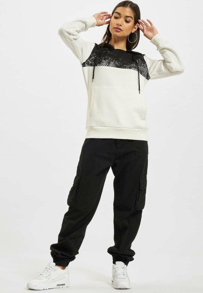 Sudadera blanca con parte superior de patrón negro, capucha con cordón y bolsillo frontal; combinada con pantalones cargo negros y zapatillas blancas.